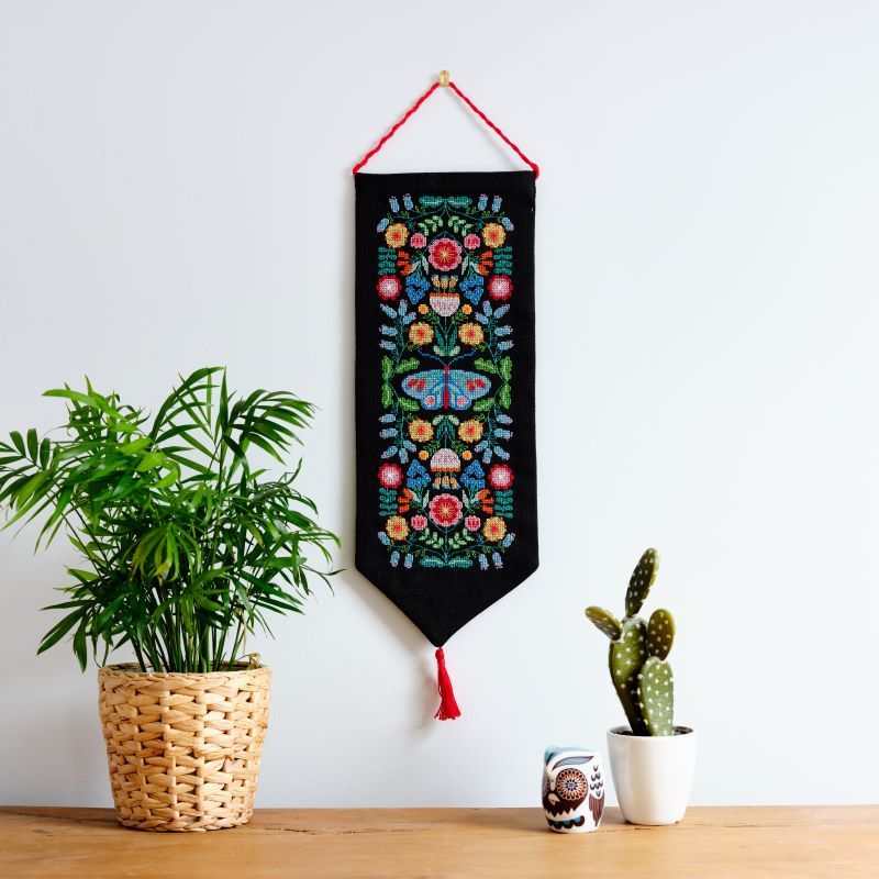 A cross stitch kit depicting a colorful floral pattern on black Aida cloth, displayed over a white background with a red hanging ribbon.