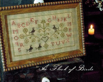 Framed cross-stitched sampler with alphabet and decorative elements, lit by a candle. A flock of birds by Blackbird design.