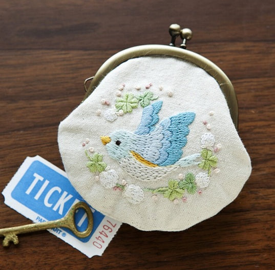 A professional-grade Japanese embroidery and pouch assembly kit by Chicchi, titled 'Bird and White Clover Coin Purse.' The high-definition package showcases a whimsical, boutique design of a stylized songbird nestled among delicate, textured white clover flowers and leaves. The kit is an authentic, artisanal package containing high-quality background fabric, a full-size purse template, premium pre-sorted embroidery threads, and a classic metal kiss-lock purse snap frame. The instruction booklet features det