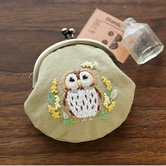 A professional-grade Japanese embroidery and pouch assembly kit by Chicchi, titled 'Owl and Mimosa Coin Purse.' The high-definition package showcases a whimsical, boutique design of a stylized owl nestled among vibrant, textured mimosa flowers. The kit is an authentic, artisanal package containing high-quality background fabric, a full-size purse template, premium pre-sorted embroidery threads, and a classic metal kiss-lock purse snap frame. The instruction booklet features detailed technical illustrations 