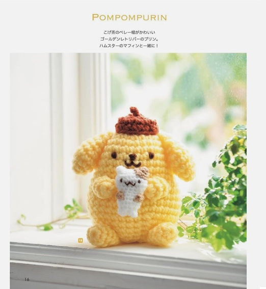 Crocheted yellow pompompurin with a red hat on a windowsill with greenery.