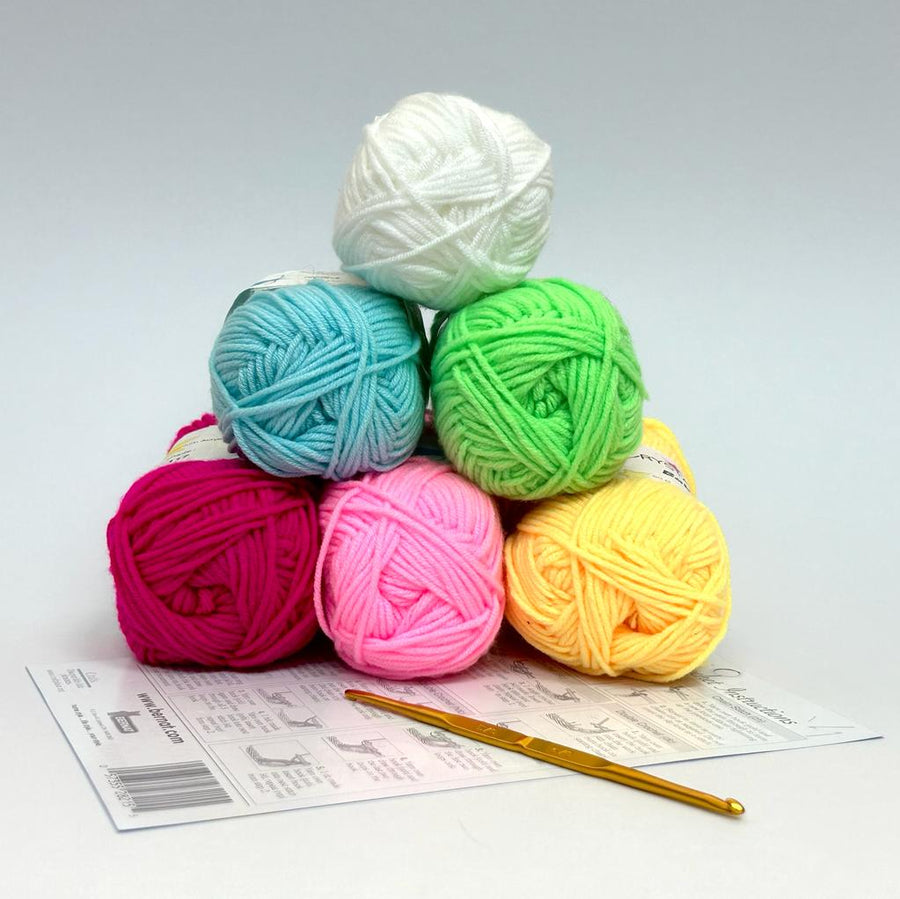 Six skeins of colorful acrylic yarn in pink, blue, green, yellow, and white, stacked together with a crochet hook lying on a piece of paper.