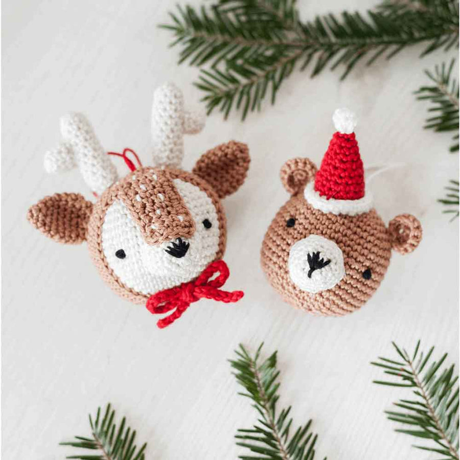 Two amigurumi crochet toys, a reindeer head with a red bow and a teddy bear wearing a Santa's red hat, made in different colors, placed on a white surface with green leaves in the background.