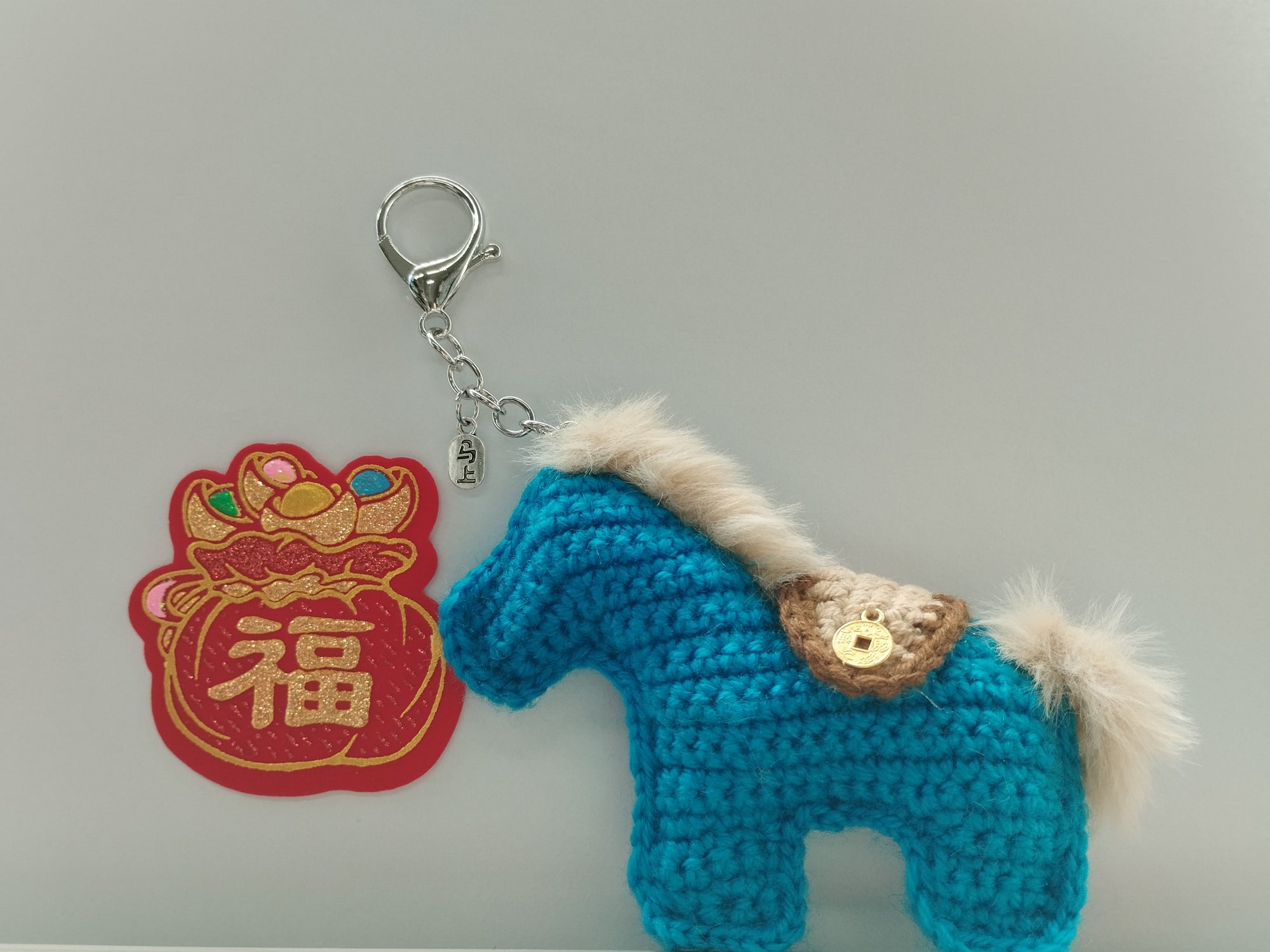 Blue crochet horse keychain with a red and gold decorative patch on a gray background