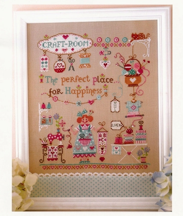 A professional cross-stitch chart by Cuore e Batticuore titled 'Il Posto Della Felicità.' The high-definition design features a charming girl in a cozy, cluttered craft room filled with fabric bolts, thread spools, floral arrangements, and heart motifs. The pattern includes the English verse for 'The perfect place for happiness