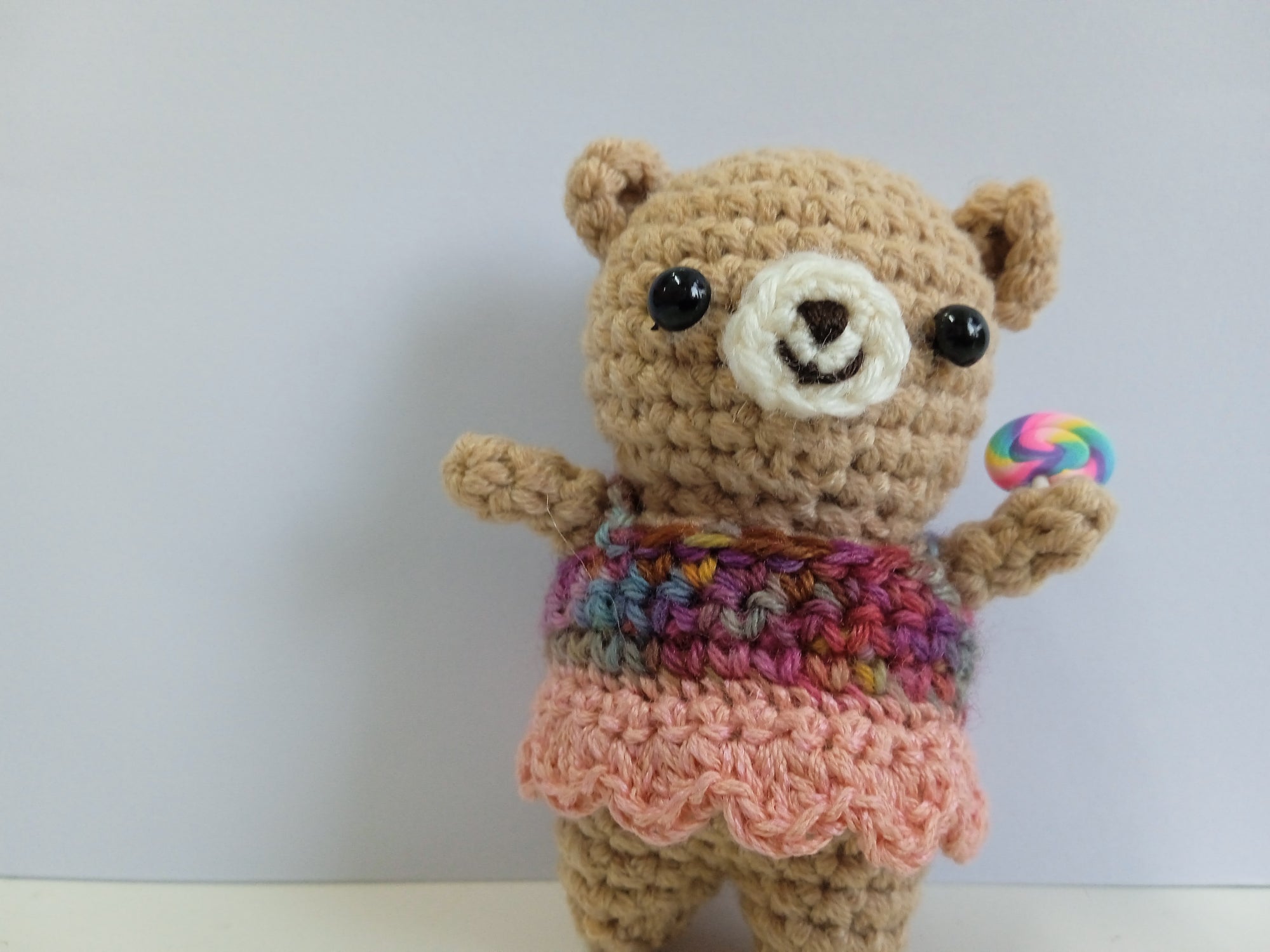 Artisan made in Singapore. Crocheted teddy bear wearing a colorful sweater and holding a lollipop against a white background. The perfect palm-sized traveling companion made from soft, durable yarn for comfort on long journeys.