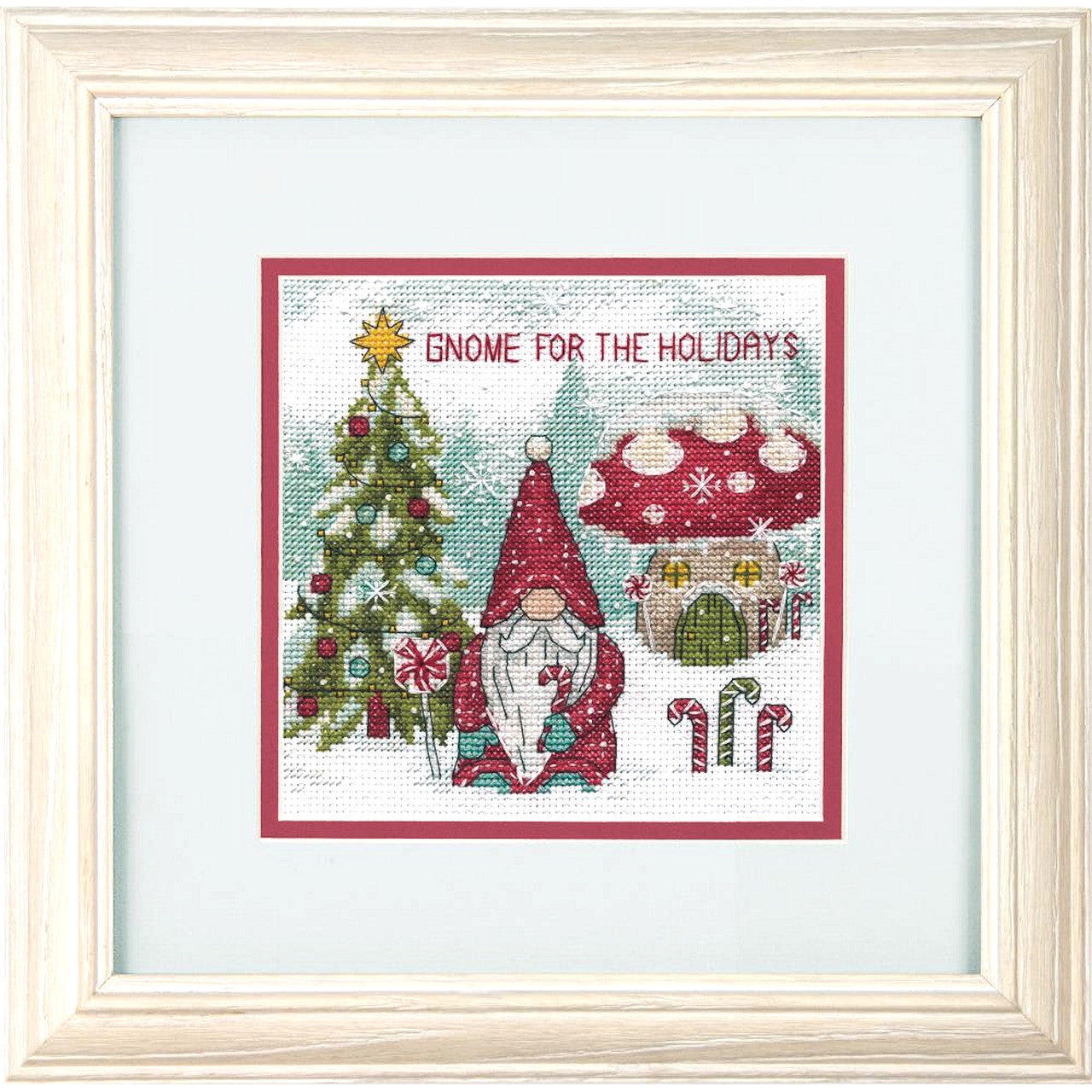 A cross-stitch depiction of a gnome with a white beard and a Santa-styled stocking cap, with a Christmas tree and holiday-themed decorations in the background. The gnome is wearing a red hat that reads 'G.