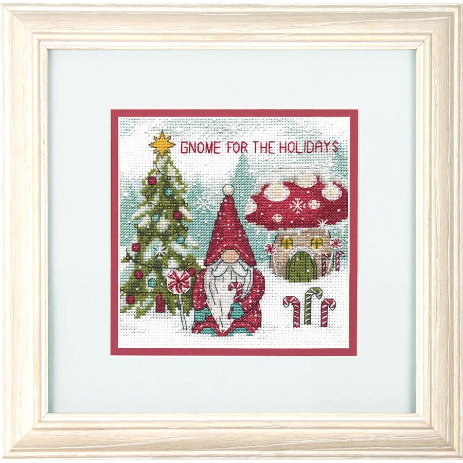 A cross-stitch depiction of a gnome with a white beard and a Santa-styled stocking cap, with a Christmas tree and holiday-themed decorations in the background. The gnome is wearing a red hat that reads 'G.