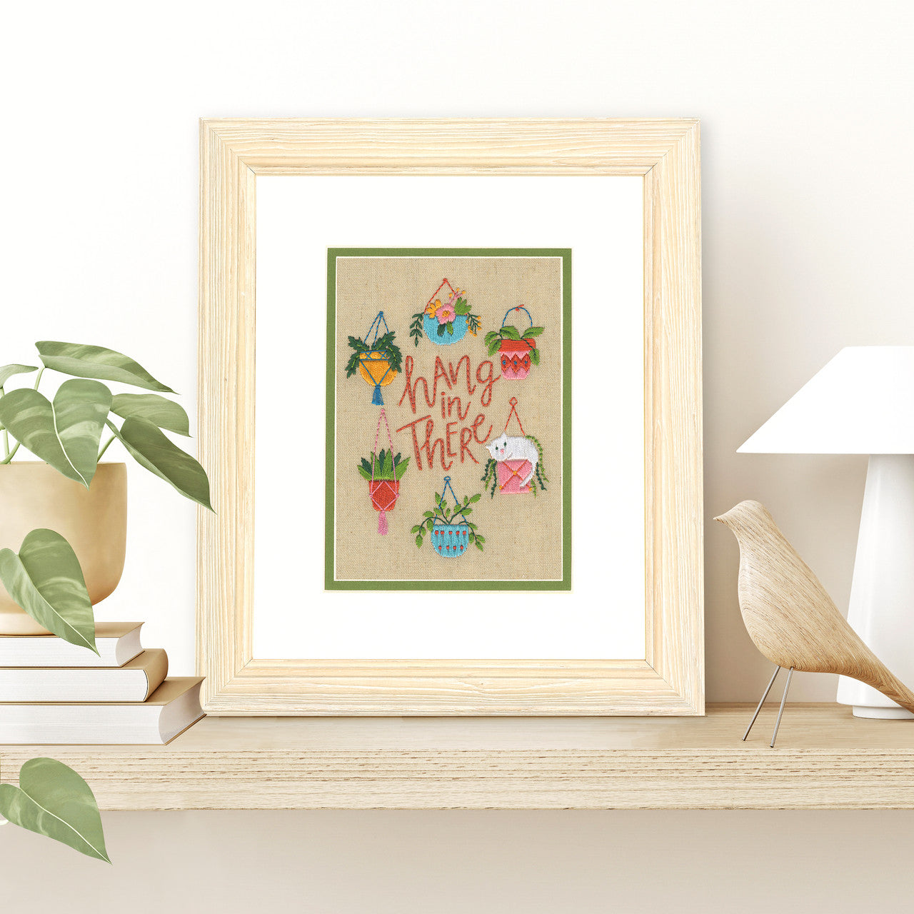 A delightful Dimensions embroidery kit featuring a small white kitten peeking out from a woven hanging basket. The kitten is surrounded by various indoor plants, including trailing vines and leafy greens in decorative pots. The encouraging phrase 'Hang in There' is centered in the design. The kit includes pre-printed linen fabric, embroidery floss, and a needle for a 7x5 inch finished piece.