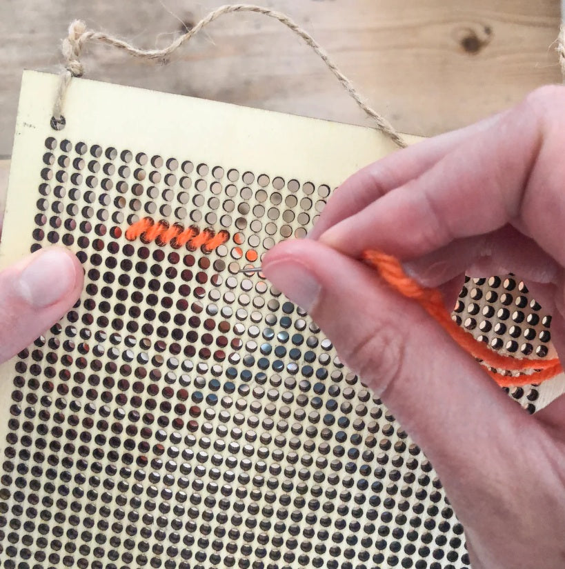 Person stitching onto a perforated fabric with orange thread on a wooden surface