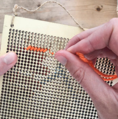 Person stitching onto a perforated fabric with orange thread on a wooden surface