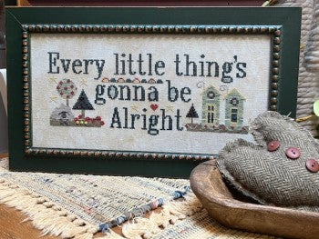Cross stitch pattern chart with the phrase 'Every little thing's gonna be alright' and accompanying visual design, displayed on a wall above a wooden bowl with a grey fabric in the foreground. Cross stitch chart for 'Every Little Thing' by Amy Bruecken featuring stylized, colorful folk-art lettering on a neutral background