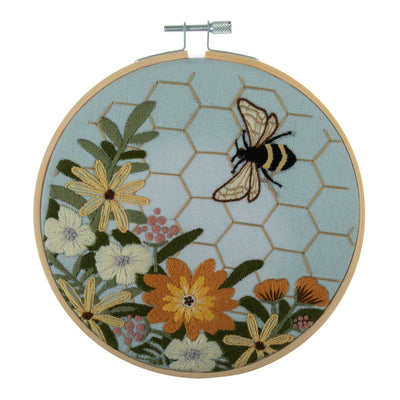 Embroidery hoop kit depicting a floral design with bee motifs and a honeycomb pattern on a light blue background, completed and framed.