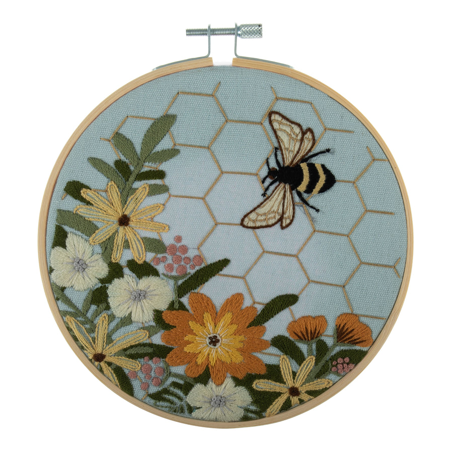Embroidery hoop kit depicting a floral design with bee motifs and a honeycomb pattern on a light blue background, completed and framed.