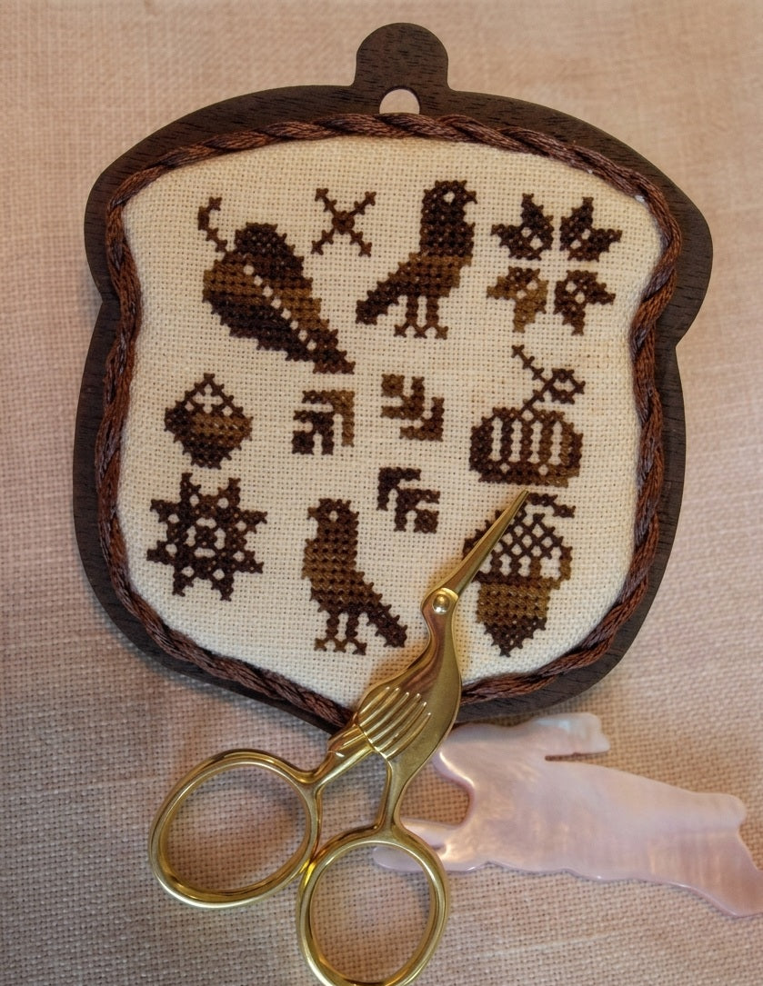 A professional-grade counted needlework chart package from Kelmscott Designs titled 'Autumn Acorn.' The high-definition product features a boutique primitive folk art cross-stitch pattern alongside an exclusive, custom hand-painted wooden acorn shape designed to be mounted onto the finished stitching. The kit is an artisanal package containing the template and detailed step-by-step structural finishing instructions.