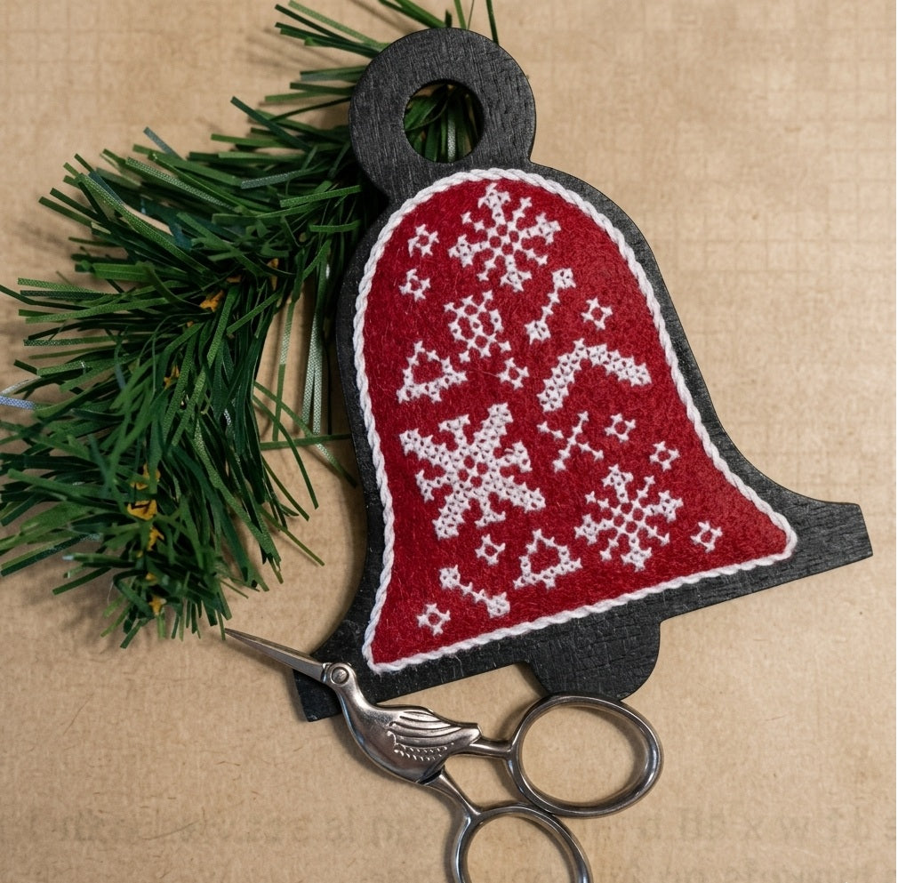 A professional needlework kit from Kelmscott Designs titled 'Christmas Bells Are Ringing.' The kit features a unique, flat-profile wooden bell form designed to be the base for an embroidered ornament. It includes a sophisticated Folk Art cross-stitch chart with traditional motifs and detailed instructions on how to finish the stitching and mount it onto the wooden bell.