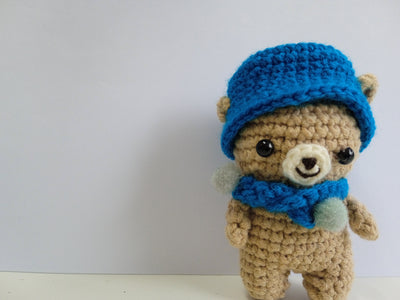 Crocheted teddy bear wearing a blue hat and scarf. Stuffed animal photography. Artisan hand made. 
The perfect palm-sized traveling companion made from soft, durable yarn for comfort on long journeys.