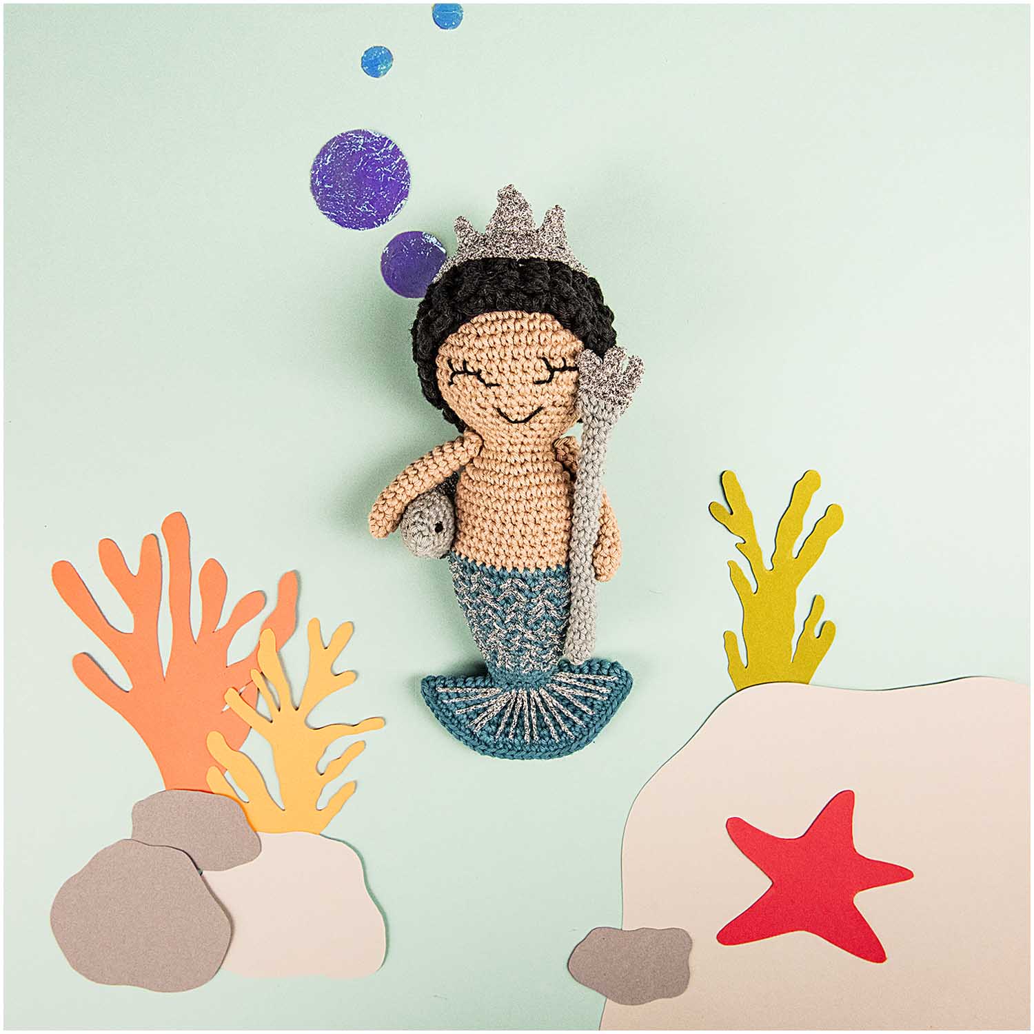 Crochet kit depicting a Neptune character with a crown, sitting on a rock, surrounded by a aquatic scene with coral and a starfish.