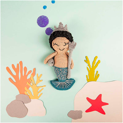 Crochet kit depicting a Neptune character with a crown, sitting on a rock, surrounded by a aquatic scene with coral and a starfish.