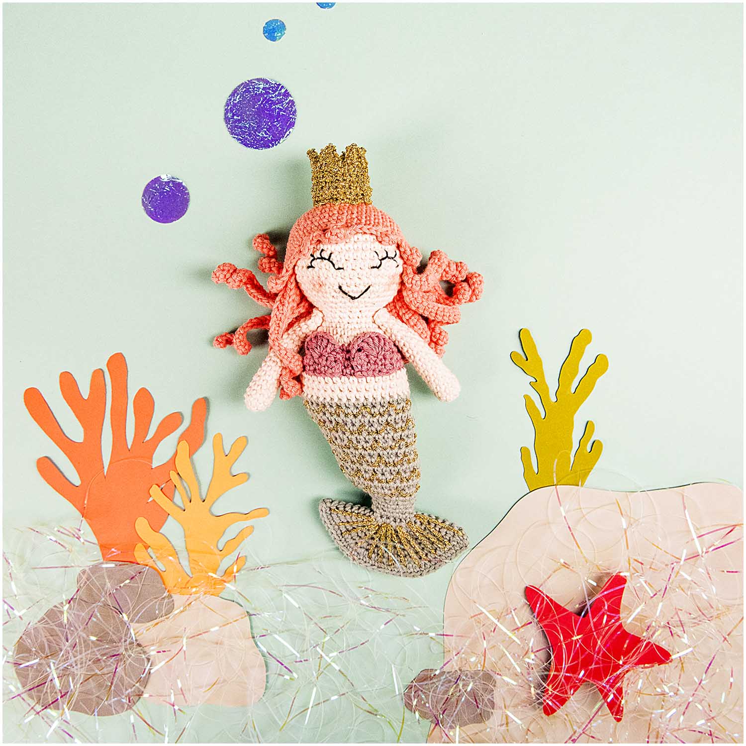 A crochet mermaid kit with pink and orange colors, including yarn, a crochet hook, and pattern instructions.