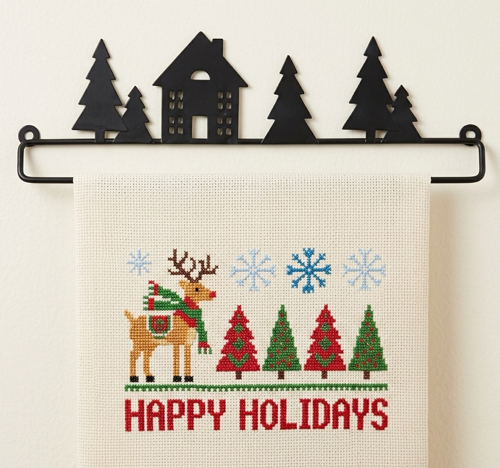 Cross-stitched towel with Christmas design featuring a reindeer, trees, and 'Happy Holidays' text, hanging on a black metal rack.