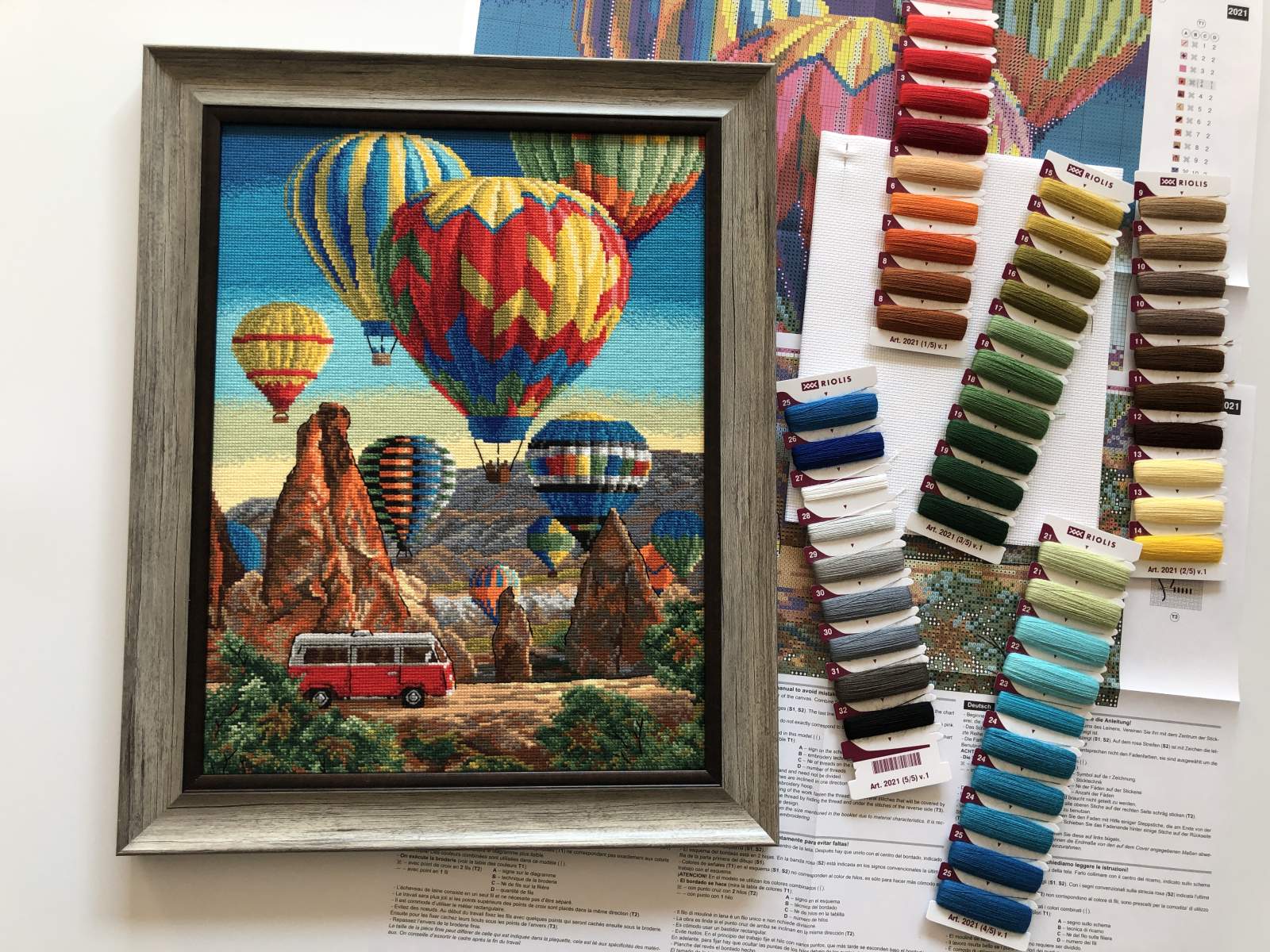 Cross stitch kit depicting colorful hot-air balloons against a Cappadocia backdrop, with a frame and matting excluded. The kit includes fabric and thread samples. Riolis Cappadocia cross stitch kit featuring a cluster of colorful hot air balloons floating over the unique 'fairy chimney' rock formations of Turkey.