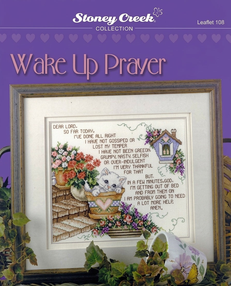 A professional-grade cross-stitch chart by Stoney Creek titled 'Wake Up Prayer.' The high-definition design features a humorous and soulful prayer about the challenges of staying patient and kind once the day truly begins. The text is bordered by whimsical, boutique-style illustrations of morning-themed items and delicate floral accents.