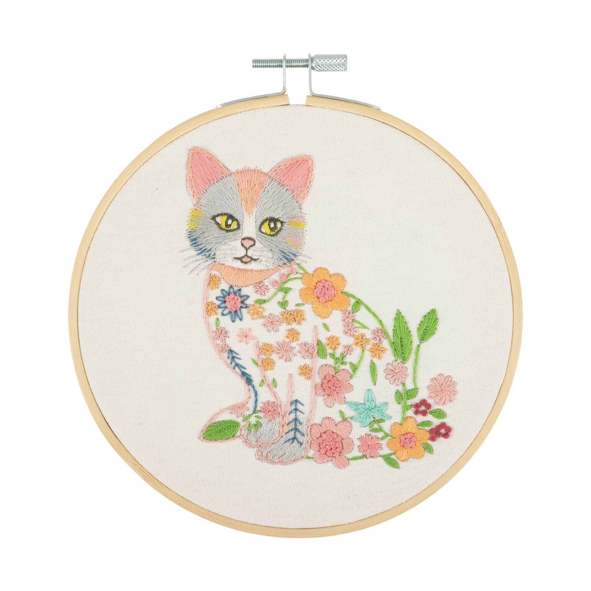 Trimits Cat Embroidery Hoop Kit featuring a charming  white cat design with simple, elegant floral accents on a neutral fabric background.
Embroidery hoop kit depicting a cat with floral patterns on a beige background, completed and framed.