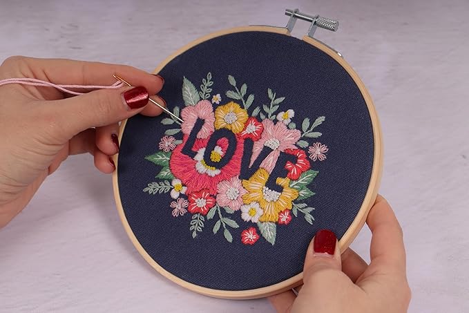 Beginner-friendly Trimits Love embroidery kit with pre-printed floral design, 15cm wooden hoop, and easy-to-follow instructions for modern needlecraft.
A person is working on an embroidery hoop with a floral design and the word 'love' in the center, using a needle and thread.
