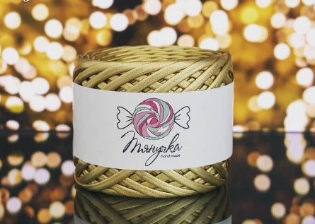 A spool of metallic gold yarn with a label that has a heart and wings graphic on it, set against a blurred bokeh background.