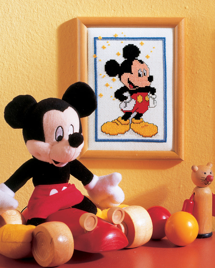 Plush toy resembling a cartoon character next to a framed cross-stitch pattern of the same character on a yellow wall.
