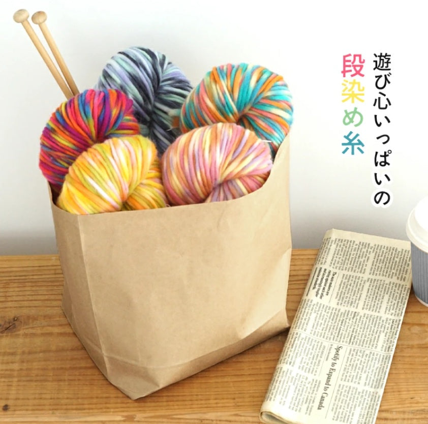 Colorful variegated yarn balls suitable for knitting and croceheing in a brown paper bag on a wooden surface with knitting needles and a newspaper. yarn made in japan