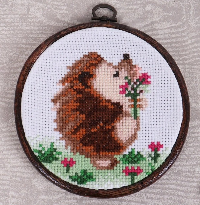 Cross-stitched hedgehog holding flowers in a wooden hoop on a textured surface