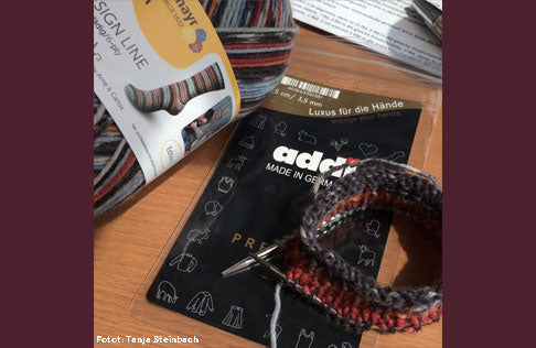 Circular knitting needles with a gold cord connecting the tips, alongside a package with knitting yarn.