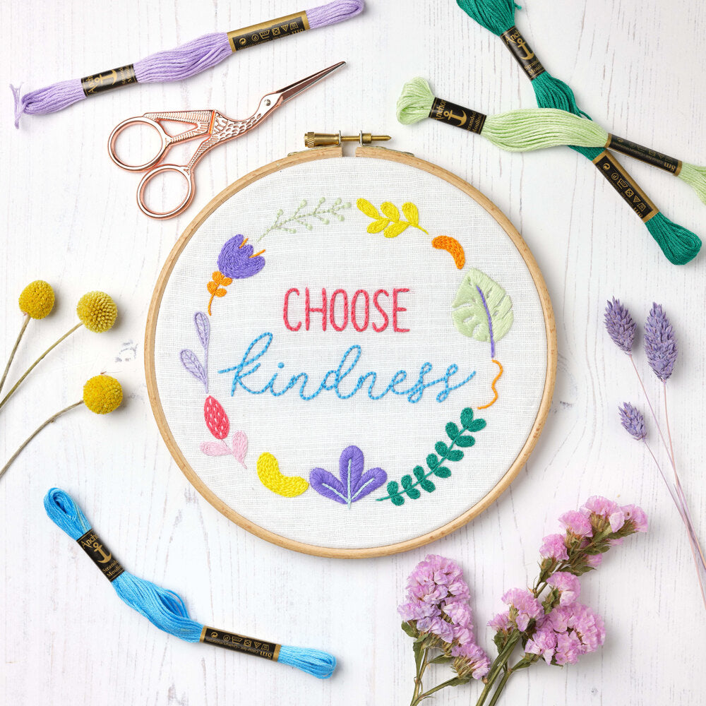 An Anchor Essentials freestyle embroidery kit featuring the 'Choose Kindness' design. The image shows a contemporary floral arrangement with modern leaf shapes and blooms surrounding the central text. The kit includes white cotton fabric with a pre-printed outline, vibrant Anchor stranded cotton threads, and all necessary tools for a 20cm x 20cm finished hoop art