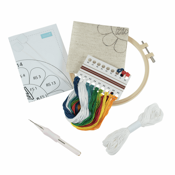 A complete "Be Kind" punch needle kit by Trimits, featuring a pre-printed fabric panel, an embroidery hoop, colorful yarn, and a punch needle tool for a textured DIY wall hanging.