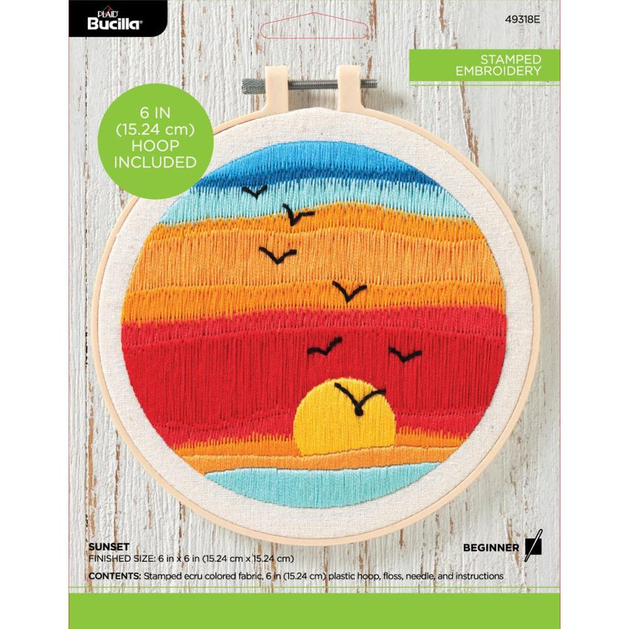 A beginner-friendly Bucilla stamped embroidery kit featuring a scenic sunset design. The kit includes a 6-inch round plastic hoop, a white broadcloth fabric pre-stamped with the sunset pattern, vibrant cotton embroidery floss in warm oranges and pinks, a needle, and easy-to-follow instructions