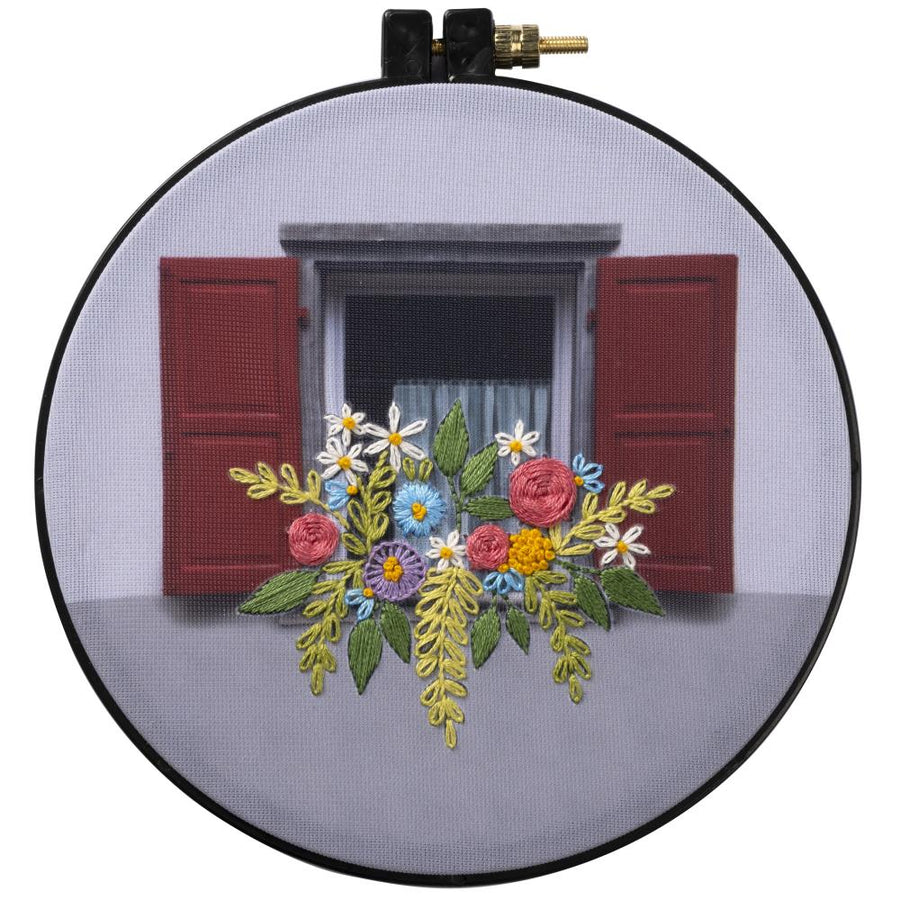 Round embroidery kit depicting a flower box scene with a red door and white flowers, finished needlework presented on a 6-inch plastic hoop.

A beginner-friendly Bucilla stamped embroidery kit featuring the 'Flower Box' design. The kit includes a photographic stamped polyester broadcloth with the floral design clearly marked, colorful cotton embroidery floss, an embroidery needle, and a 6-inch blue plastic hoop that serves as both your workspace and your final display frame