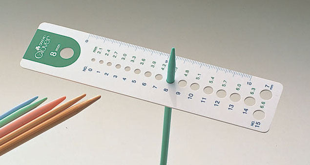 A professional Clover brand knitting needle gauge made in Japan. The tool is a durable, flat rectangular plate featuring a series of precision-drilled holes for sizing knitting needles. Each hole is marked with high-definition metric and US sizes. One side of the gauge includes a 10cm ruler for quick measurements. The iconic Clover logo is embossed on the high-quality surface, identifying it as a premium boutique tool.