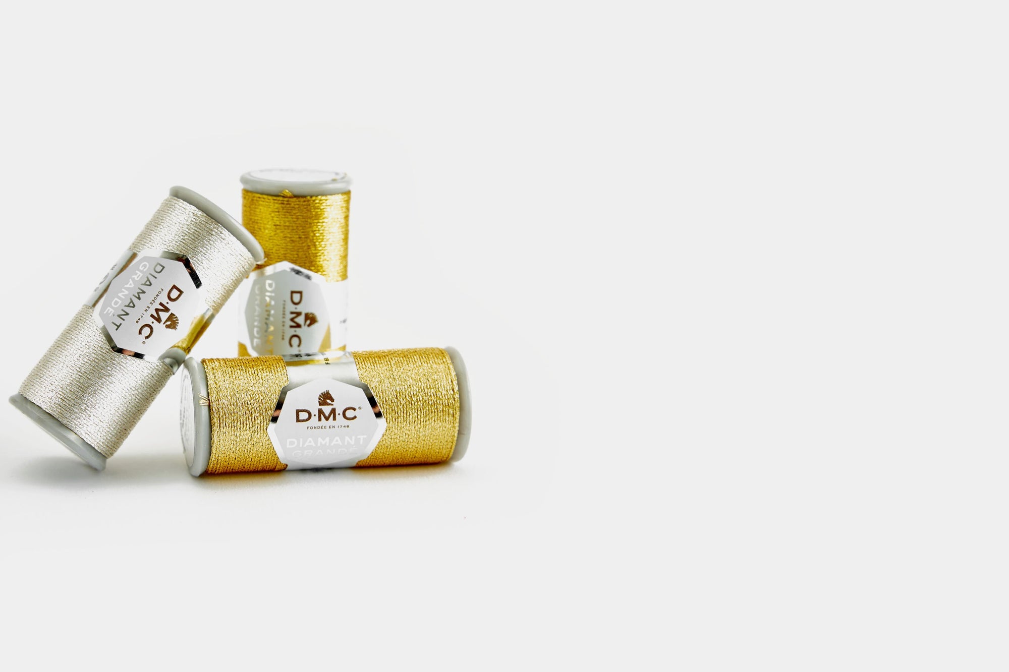 Three spools of DMC Diamant Grandé metallic thread, with two gold-colored spools and one silver-colored spool, arranged diagonally on a white background.