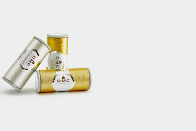 Three spools of DMC Diamant Grandé metallic thread, with two gold-colored spools and one silver-colored spool, arranged diagonally on a white background.
