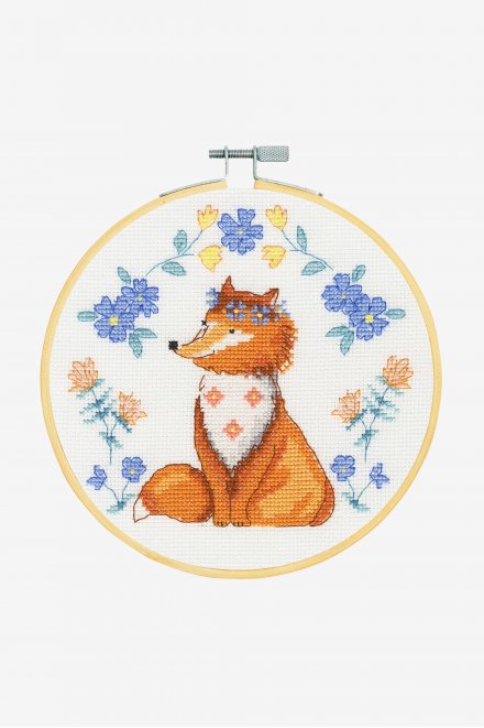 A vibrant DMC counted cross-stitch kit titled 'Folk Fox.' The design features a centered orange fox decorated with symmetrical folk-art floral patterns in teal, cream, and red. The kit is comprehensive, including 14-count white Aida fabric, a 7-inch bamboo embroidery hoop, premium DMC embroidery floss, a tapestry needle, and full instructions