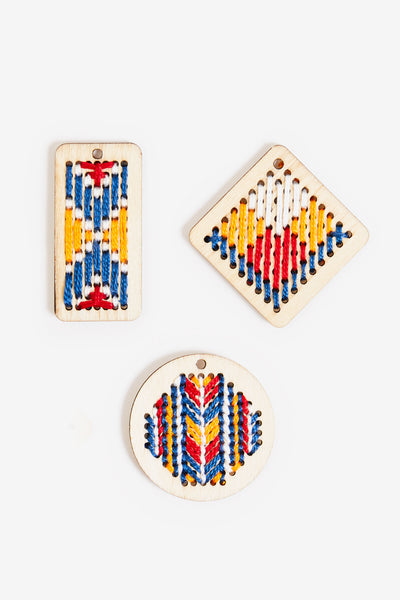 A set of three high-quality wooden jewelry pendants by DMC. The pack includes three distinct geometric shapes: one circular, one rectangular, and one diamond-shaped pendant. Each pendant features a laser-cut perforated grid, specifically spaced to accommodate embroidery floss or thin yarns. The natural wood finish offers a rustic, artisanal canvas for custom cross-stitch or needlepoint designs.