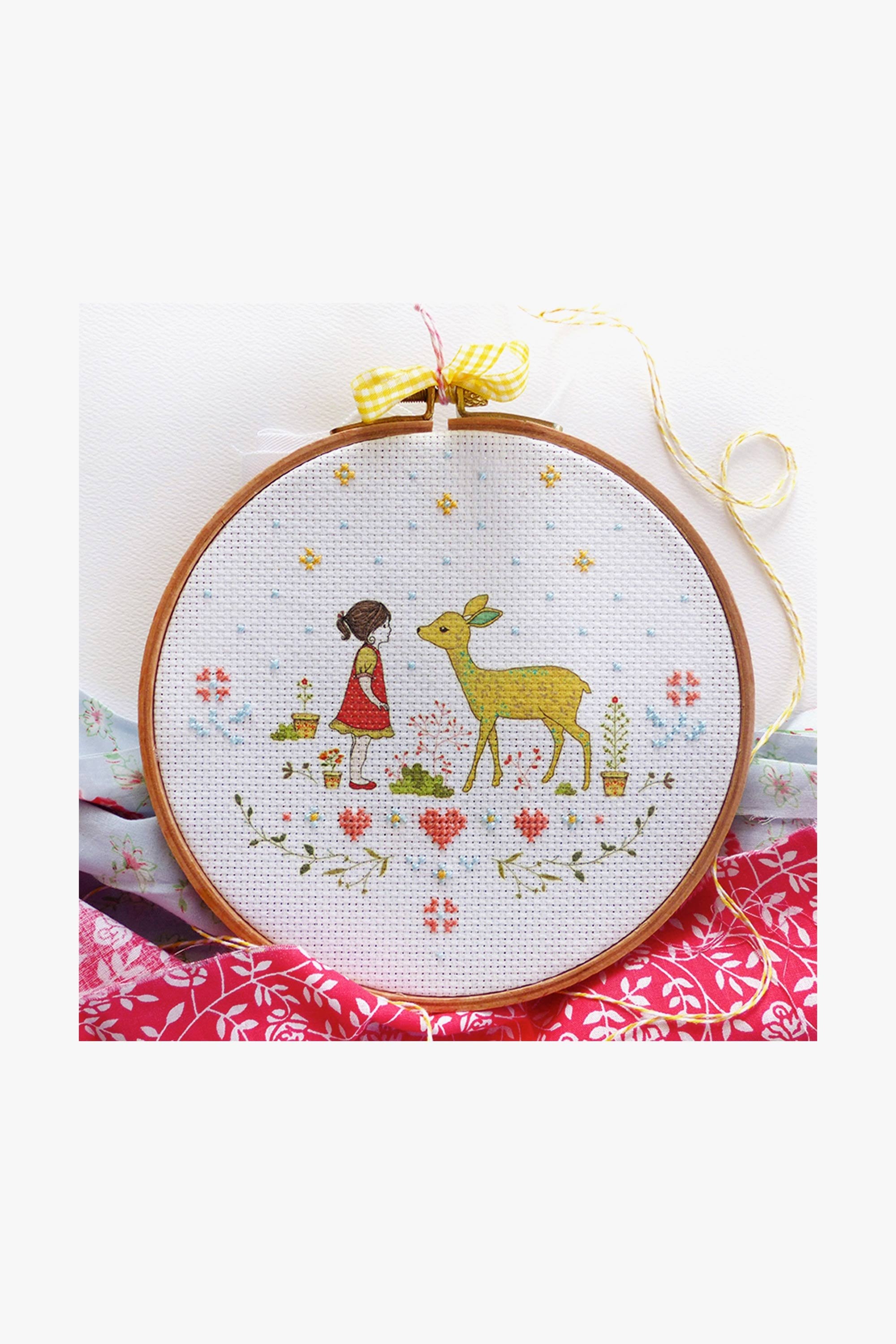 A cross stitch kit depicting a girl and a deer with a printed fabric design on 14 count Aida Cloth, accompanied by threads and a needle, displayed on a red and white patterned surface.
