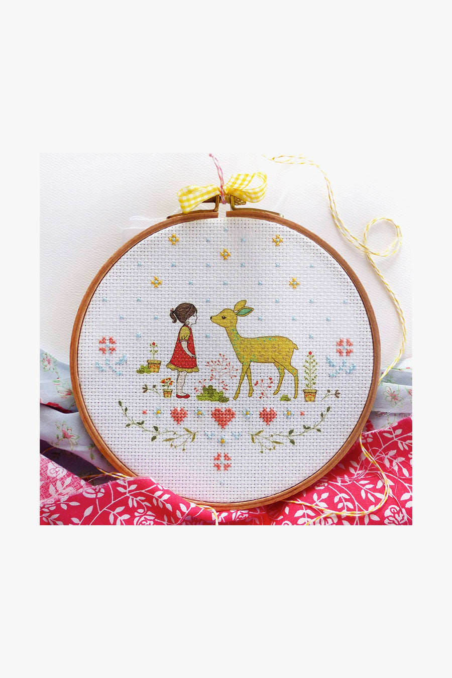 A cross stitch kit depicting a girl and a deer with a printed fabric design on 14 count Aida Cloth, accompanied by threads and a needle, displayed on a red and white patterned surface.