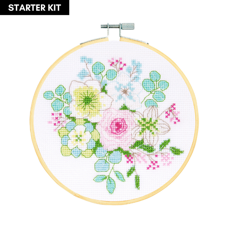 A serene and modern DMC counted cross-stitch kit titled 'Winter Bouquet.' The design features a dense, circular arrangement of winter-themed plants, including pine needles, blue-toned berries, and stylized winter flowers. The kit is all-inclusive, featuring 14-count white Aida fabric, a 6-inch bamboo embroidery hoop, premium DMC embroidery floss, a tapestry needle, and complete instructions