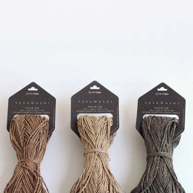 Three skeins of Daruma Sasawashi yarn in beige, black, and brown colors, displayed against a white background.