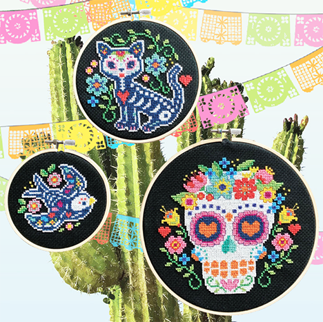 A cross-stitch pattern chart featuring three separate 'Día de los Muertos' inspired designs. The trio includes a bird, a cat, and a sugar skull, all decorated with vibrant flowers, symmetrical leaf motifs, and colorful geometric patterns. The chart provides clear symbols and a color legend for creating these folk-art-style embroideries.