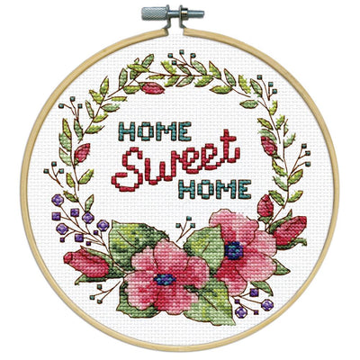 A beginner-friendly Design Works counted cross-stitch kit featuring a 'Floral Wreath' design. The image showcases a classic circular garland of pink flowers with green leaves. The kit includes 11-count white Aida fabric, a pre-sorted 100% cotton thread palette, a needle, and an 8-inch circular hoop for stitching and display.