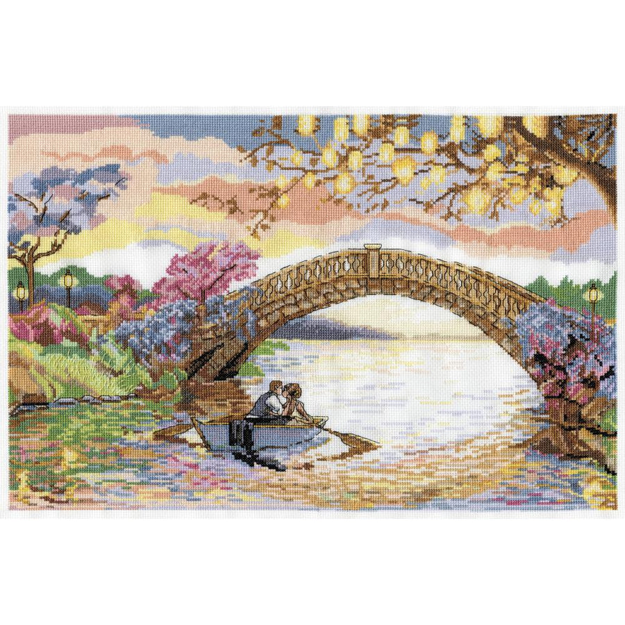 A large-scale Design Works counted cross-stitch kit titled 'Forever Kiss.' The artwork depicts a romantic scene of a couple kissing in a rowboat on a tranquil river, framed by a stone bridge and lush flowers under a glowing sunset. The kit is shown with 14-count white Aida fabric, a pre-sorted thread palette of 6-strand cotton floss, and a detailed pattern chart